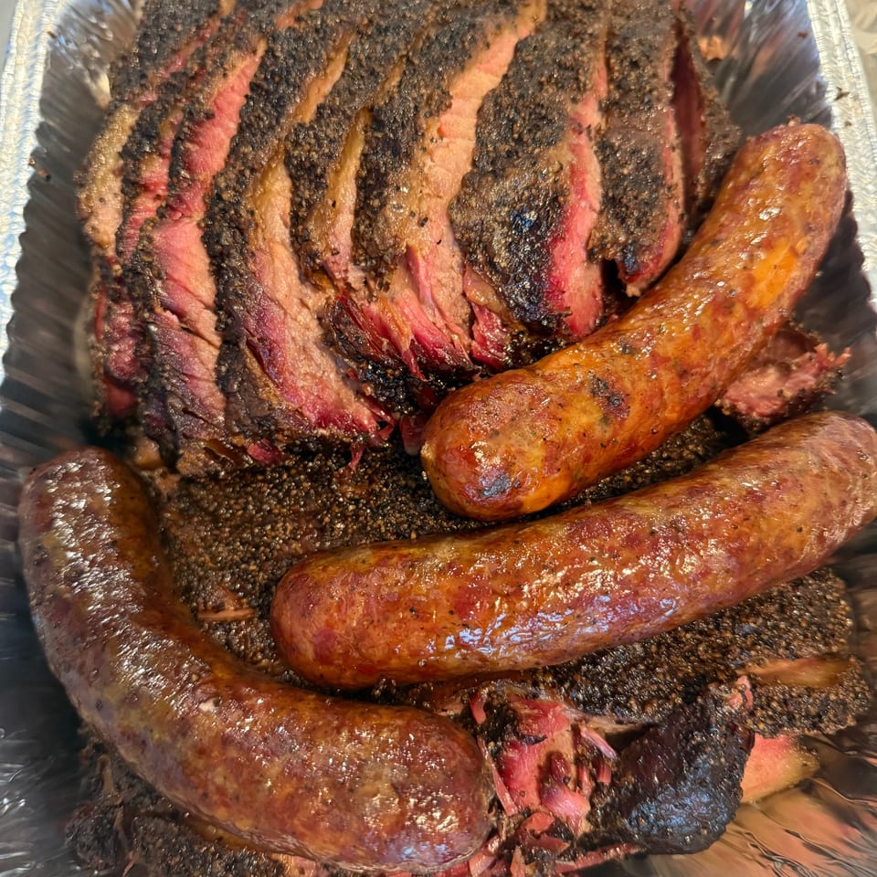 Sliced brisket with a dark bark and two large sausages in a foil pan.